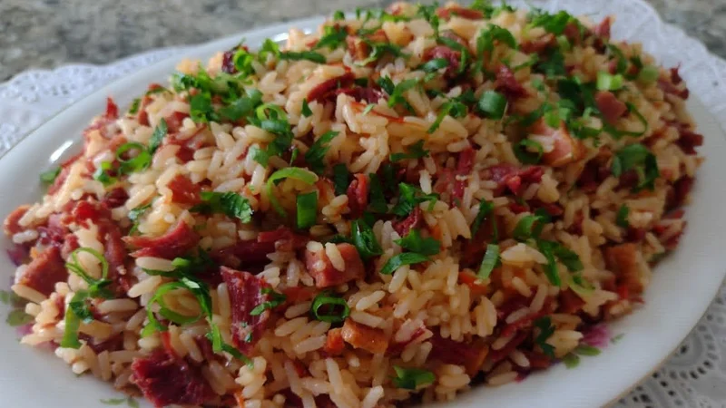 # **Arroz de Carreteiro na Panela de Cerâmica**: Receita Tradicional Gaúcha que Derrete na Boca O aroma inconfundível do **arroz de carreteiro na panela de cerâmica** me transporta diretamente para os domingos na fazenda do meu avô, no interior do Rio Grande do Sul. Lembro-me vividamente de como ele preparava este prato icônico da culinária gaúcha, sempre insistindo que só ficava perfeito quando feito na sua velha panela de barro. Hoje entendo o porquê: a cerâmica tem o poder mágico de transformar ingredientes simples em uma explosão de sabores que se harmonizam perfeitamente. Esta **receita de arroz de carreteiro** é muito mais que um prato; é uma herança cultural que carrega consigo histórias de tropeiros, viagens longas pelos pampas e a necessidade de criar refeições saborosas com poucos ingredientes. A técnica de preparo **na panela de cerâmica** não é apenas tradicionalismo, é ciência culinária aplicada: o cozimento uniforme e lento permite que cada grão de arroz absorva completamente o sabor da carne seca, criando uma textura cremosa por fora e soltinha por dentro. Descobri ao longo dos anos que muitas pessoas têm dificuldade em reproduzir o verdadeiro sabor do carreteiro, especialmente quando usam panelas comuns. A diferença está nos detalhes que vou compartilhar com vocês nesta receita testada e aprovada por três gerações da minha família. ## Por Que o **Arroz de Carreteiro na Panela de Cerâmica** é Especial A magia do **arroz de carreteiro na cerâmica** está na forma como este utensílio milenar potencializa cada ingrediente. Diferente das panelas de alumínio ou aço inoxidável, a cerâmica distribui o calor de maneira gradual e uniforme, evitando que o arroz grude no fundo e permitindo que os sabores se desenvolvam lentamente. Quando preparo meu **carreteiro na panela de barro**, posso literalmente sentir a diferença já nos primeiros minutos de cozimento. A carne seca libera seus óleos naturais de forma mais completa, o tempero se integra melhor aos grãos, e o resultado final tem aquela consistência cremosa que só conseguimos com este método tradicional. Uma das grandes vantagens da **receita de arroz de carreteiro** preparada desta forma é a capacidade da cerâmica de manter a temperatura por muito mais tempo. Isso significa que o prato continua "cozinhando" mesmo após retirarmos do fogo, desenvolvendo sabores ainda mais complexos. Minha avó costumava dizer que "o carreteiro de panela de barro fica bom, mas de panela de ferro fica sem alma" - e ela estava absolutamente certa. Além dos benefícios gustativos, cozinhar **arroz de carreteiro na cerâmica** é também uma escolha mais saudável. Este material não libera substâncias tóxicas mesmo em altas temperaturas, ao contrário de alguns revestimentos antiaderentes modernos. A porosidade natural da cerâmica também permite que os alimentos "respirem" durante o cozimento, resultando em texturas mais interessantes. Para famílias que apreciam a culinária tradicional, este método de preparo se torna quase um ritual. O tempo necessário para aquecer a panela, o cuidado na regulagem do fogo, a paciência para esperar o ponto ideal - tudo isso faz parte da experiência gastronômica que nossos antepassados nos legaram. ## Ingredientes para o Melhor **Arroz de Carreteiro na Panela de Cerâmica** ### Ingredientes Principais (serve 6 pessoas generosas): - **500g de carne seca** (charque) - escolha peças com boa distribuição de gordura - **2 xícaras de arroz agulhinha** (400g) - prefira arroz tipo 1, de grãos longos - **1 cebola grande** (cerca de 200g) cortada em cubos médios - **4 dentes de alho** bem picados - **2 tomates maduros médios** sem pele, cortados em cubos - **1 pimentão vermelho** cortado em tiras finas - **3 colheres de sopa de óleo** ou banha de porco - **4 xícaras de água quente** (pode precisar de mais) - **Sal a gosto** (cuidado, a carne seca já é salgada) - **Pimenta-do-reino moída na hora** - **2 folhas de louro** - **1 maço de cebolinha verde** picada - **Salsinha fresca** para finalizar ### Dicas de Escolha dos Ingredientes: A qualidade da **carne seca para arroz de carreteiro** faz toda a diferença no resultado final. Procure peças que não estejam ressecadas demais, com uma cor avermelhada uniforme e que tenham pequenas veias de gordura - isso garantirá sabor e suculência ao prato. Se a carne estiver muito salgada, deixe-a de molho por algumas horas antes do preparo. Para o arroz, sempre uso o tipo agulhinha (grãos longos) pois ele absorve melhor os sabores sem se desfazer durante o cozimento na cerâmica. O arroz parboilizado também funciona bem, mas requer um pouco mais de líquido. A escolha dos temperos é crucial: alho e cebola devem estar bem frescos, os tomates precisam estar maduros mas firmes (se estiverem muito moles, podem desfazer durante o cozimento), e o pimentão adiciona cor e um leve dulçor que equilibra o sal da carne seca. ### Substitutos e Variações: Se não encontrar carne seca de boa qualidade, pode usar **carne de sol** (menos salgada) ou até mesmo **jerked beef** - neste caso, ajuste a quantidade de sal. Para uma versão mais suave, experimente misturar metade carne seca e metade linguiça calabresa defumada. Vegetarianos podem adaptar usando cogumelos portobello desidratados e reintratados, que conferem um sabor umami interessante, embora obviamente diferente do original. ## Modo de Preparo do **Arroz de Carreteiro na Panela de Cerâmica** ### Preparação Prévia (Mise en Place): Antes de iniciar o preparo do **arroz de carreteiro na cerâmica**, organize todos os ingredientes. Corte a carne seca em cubos de aproximadamente 2cm, deixe o arroz lavado e escorrido, tenha todos os temperos picados e a água já aquecida. Esta organização é fundamental pois o processo não permite pausas longas. ### Passo 1: Tratamento da Carne Seca (15 minutos) Coloque a carne seca em uma panela com água fervente e deixe cozinhar por 10 minutos. Este processo remove o excesso de sal e amacia as fibras. Escorra e reserve. Se preferir um sabor mais intenso, pule esta etapa, mas ajuste o sal do prato. ### Passo 2: Aquecimento da Panela de Cerâmica (5 minutos) Aqueça sua **panela de cerâmica** em fogo baixo por alguns minutos antes de adicionar qualquer ingrediente. Este passo é crucial para evitar choque térmico que pode trincar a cerâmica. A panela está no ponto ideal quando você consegue manter a mão a 10cm da superfície por apenas 3 segundos. ### Passo 3: Refogado Inicial (8-10 minutos) Adicione o óleo ou banha na panela aquecida e refogue a carne seca em fogo médio até dourar levemente. A gordura liberada pela carne será fundamental para o sabor do **arroz de carreteiro**. Mexa ocasionalmente para dourar uniformemente. ### Passo 4: Sofrito dos Temperos (5 minutos) Adicione a cebola picada e refogue até ficar transparente. Junte o alho e refogue por mais 1 minuto, sempre mexendo para não queimar. O segredo aqui é deixar os temperos liberarem todos os aromas antes de prosseguir. ### Passo 5: Incorporação dos Vegetais (7 minutos) Adicione o tomate picado e o pimentão. Cozinhe até o tomate começar a se desfazer e formar um molho encorpado. Tempere com as folhas de louro, pimenta-do-reino e uma pitada de sal (lembre-se que a carne já é salgada). ### Passo 6: Tostagem do Arroz (5 minutos) Este é o passo mais importante para um **arroz de carreteiro na panela de cerâmica** perfeito. Adicione o arroz lavado e escorrido, misture bem com os demais ingredientes e deixe "torrar" levemente. Você saberá que está no ponto quando os grãos começarem a estalar suavemente e ficarem translúcidos. ### Passo 7: Adição do Líquido (2 minutos) Adicione a água quente de uma só vez - nunca água fria, pois pode trincar a cerâmica. A proporção ideal é de 2 partes de líquido para 1 de arroz, mas na cerâmica geralmente preciso de um pouco mais devido à evaporação mais lenta. ### Passo 8: Cozimento Principal (25-30 minutos) Após a fervura inicial, reduza o fogo para bem baixo, tampe a panela e deixe cozinhar. A **cerâmica para arroz de carreteiro** trabalha de forma diferente: o calor continua distribuído mesmo em fogo baixo. Resista à tentação de mexer - deixe a panela fazer seu trabalho. ### Passo 9: Descanso e Finalização (10 minutos) Quando a água secar completamente, retire do fogo e deixe descansar tampado por 10 minutos. Este descanso permite que o vapor termine o cozimento e que os sabores se assentem. Apenas então destampe, misture delicadamente e finalize com cebolinha e salsinha. ### Passo 10: Ponto Final e Apresentação (5 minutos) O **arroz de carreteiro** está pronto quando cada grão está soltinho mas cremoso, com a carne bem distribuída e um aroma irresistível. Prove e ajuste o tempero se necessário, lembrando que o sabor se intensifica após o descanso. ## Dicas e Variações do **Arroz de Carreteiro na Panela de Cerâmica** Durante anos aperfeiçoando esta **receita de arroz de carreteiro**, desenvolvi algumas técnicas que fazem toda a diferença no resultado final. A primeira e mais importante é sobre o controle da temperatura: a cerâmica retém calor por muito tempo, então sempre reduzo o fogo mais cedo do que faria com panelas convencionais. ### Variações Testadas e Aprovadas: **Carreteiro com Linguiça:** Substitua metade da carne seca por linguiça calabresa cortada em rodelas. Doure primeiro a linguiça para liberar a gordura, depois siga o processo normal. Esta versão fica especialmente saborosa **na panela de cerâmica**. **Versão com Bacon:** Para um toque mais defumado, adicione 100g de bacon cortado em cubinhos junto com a carne seca. O resultado é um **arroz de carreteiro** com camadas de sabor ainda mais complexas. **Carreteiro Vegetal:** Experimente adicionar cenoura cortada em cubinhos pequenos e vagem picada junto com o pimentão. Estes vegetais ficam perfeitos quando cozidos lentamente **na cerâmica**. **Versão Picante:** Para quem aprecia pratos mais temperados, adicione uma pimenta dedo-de-moça sem sementes, cortada em anéis finos, junto com os temperos iniciais. ### Segredos da Vovó: Minha avó sempre dizia que o **arroz de carreteiro na panela de barro** devia "cantar" quando estava pronto - esse som suave de crepitação indica que chegou ao ponto ideal. Ela também adicionava uma colher de café de açúcar no refogado inicial, que ajuda a caramelizar os temperos e dar uma cor mais bonita ao prato. Outro truque que aprendi é salgar a água do cozimento da carne seca com uma folha de louro - isso remove o sal em excesso mas mantém o sabor característico. ### Acompanhamentos Perfeitos: O **carreteiro de panela de cerâmica** combina perfeitamente com farofa de banana, vinagrete de cebola roxa, couve refogada ou uma salada simples de alface com tomate. Para bebidas, um mate gelado ou uma cerveja bem gelada são acompanhamentos clássicos. ### Dicas de Apresentação: Sirva o **arroz de carreteiro** diretamente da panela de cerâmica para manter a temperatura. Se quiser uma apresentação mais sofisticada, transfira para uma travessa aquecida e decore com rodelas finas de tomate e raminhos de salsinha fresca. ## Como Conservar e Reaproveitar o **Arroz de Carreteiro** Uma das grandes vantagens do **arroz de carreteiro na panela de cerâmica** é que ele fica ainda mais saboroso no dia seguinte. A cerâmica continua desenvolvendo os sabores mesmo após o resfriamento, e quando reaquecemos, obtemos um prato com notas ainda mais complexas. ### Armazenamento Correto: Deixe o **carreteiro** esfriar completamente na própria panela antes de transferir para recipientes. Na geladeira, mantém-se perfeito por até 4 dias em potes bem vedados. Nunca deixe na panela de cerâmica dentro da geladeira, pois a umidade pode ser prejudicial ao material. ### Formas de Reaquecer: Para reaquecer, prefira sempre uma panela com um fio de óleo e algumas colheres de água quente. Mexe delicadamente até aquecer uniformemente. Microondas também funciona, mas perde um pouco da textura original. ### Receitas de Reaproveitamento: Com o **arroz de carreteiro** que sobrou, você pode preparar deliciosos bolinhos: misture com um ovo batido, forme bolinhas e frite em óleo quente. Outra opção é usar como recheio para pastéis ou empadas - fica sensacional! Também gosto de preparar um "carreteiro refogado": aqueço o arroz com ovos mexidos, cebolinha fresca e uma pitada de pimenta caiena. Vira um prato completamente novo e igualmente delicioso. ### Congelamento: O **arroz de carreteiro** pode ser congelado por até 3 meses em porções individuais. Descongele na geladeira overnight e reaqueça com cuidado, adicionando um pouco de água se necessário para recuperar a cremosidade. ## Benefícios Nutricionais e Históricos do **Arroz de Carreteiro** Este prato tradicional gaúcho nasceu da necessidade dos tropeiros de preparar refeições nutritivas e saborosas durante as longas viagens pelos pampas. A combinação de **arroz e carne seca** fornece proteínas completas, carboidratos complexos e a energia necessária para jornadas extenuantes. ### Valor Nutricional: Uma porção de **arroz de carreteiro na panela de cerâmica** (aproximadamente 200g) fornece cerca de 320 calorias, 18g de proteínas, 45g de carboidratos e 8g de lipídios. A carne seca é rica em ferro, zinco e vitaminas do complexo B, enquanto o arroz oferece energia sustentável. Os vegetais adicionados (cebola, tomate, pimentão) contribuem com vitaminas C, A e antioxidantes importantes para o sistema imunológico. O método de preparo **na cerâmica** preserva melhor estes nutrientes pois o cozimento é mais suave e uniforme. ### Aspecto Cultural: Preparar **arroz de carreteiro** é manter viva uma tradição que conecta gerações. Cada família gaúcha tem sua versão, seus segredos, suas histórias. Quando cozinho **na panela de cerâmica**, sinto que estou participando de um ritual ancestral que vai muito além da simples preparação de uma refeição. Esta receita representa a resourcefulness (capacidade de adaptação) do povo gaúcho, que transformou ingredientes simples e duradouros em um prato reconhecido nacionalmente. É uma demonstração de como a culinária regional pode ser sofisticada em sua simplicidade. ## Equipamentos e Utensílios para o **Arroz de Carreteiro na Cerâmica** ### Panela de Cerâmica Ideal: Para preparar **arroz de carreteiro** para 6 pessoas, use uma panela de cerâmica com capacidade mínima de 3 litros. Prefira modelos com fundo espesso e tampa que vede bem. Panelas de Minas Gerais, feitas em cerâmica vidrada, são especialmente boas para esta receita. ### Cuidados com a Panela: Antes do primeiro uso, tempere sua panela deixando-a de molho em água por 24 horas. Para limpeza, use apenas água morna e detergente suave - nunca produtos abrasivos que podem danificar a superfície. ### Utensílios Complementares: - Colher de pau para mexer (evita arranhar a cerâmica) - Faca bem afiada para cortar a carne seca - Tábua de corte dedicada para carnes - Panela auxiliar para dessalgar a carne - Escorredor para lavar o arroz ### Alternativas à Panela de Cerâmica: Se não tiver uma panela de cerâmica, pode usar panela de ferro fundido esmaltado ou até mesmo uma caçarola de barro. O importante é que seja um material que distribua o calor uniformemente e retenha temperatura. ## Erros Comuns ao Preparar **Arroz de Carreteiro na Panela de Cerâmica** Durante anos ensinando esta receita, identifiquei os erros mais frequentes que podem comprometer o resultado final do **arroz de carreteiro**. O primeiro e mais comum é não dessalgar adequadamente a carne seca, resultando em um prato excessivamente salgado. ### Problemas de Temperatura: Muita gente comete o erro de usar fogo alto demais na cerâmica, o que pode queimar o fundo do arroz enquanto a parte superior permanece crua. A **panela de cerâmica** trabalha melhor em temperaturas moderadas e constantes. ### Questões de Timing: Outro erro frequente é mexer o arroz durante o cozimento. Diferente de outros métodos, o **arroz de carreteiro na cerâmica** deve ser deixado em paz para que os grãos cozinhem uniformemente e desenvolvam a textura ideal. ### Problemas com Líquidos: Usar água fria ou adicionar líquido insuficiente são falhas comuns. A cerâmica evapora líquidos mais lentamente, mas o arroz ainda precisa da quantidade adequada para hidratar completamente. ### Como Corrigir: Se o arroz ficar salgado demais, prepare uma pequena porção de arroz branco e misture. Se ficar muito seco, adicione água quente aos poucos e deixe descansar tampado. Se grudar no fundo, não raspe - sirva por cima e use água morna para amolecer depois. ## **Arroz de Carreteiro na Panela de Cerâmica**: Uma Tradição que Vale a Pena Preservar Chegamos ao final desta jornada gastronômica, e espero que você tenha sentido não apenas vontade de preparar esta receita, mas também de compreender seu valor cultural. O **arroz de carreteiro na panela de cerâmica** é mais que comida - é memória, tradição e amor servidos à mesa. Esta receita, testada e refinada ao longo de anos, representa a essência da culinária gaúcha: ingredientes simples, técnicas tradicionais e muito sabor. Quando você preparar seu primeiro **carreteiro na cerâmica**, lembre-se de que está participando de uma tradição centenária que conecta famílias e gerações. Encorajo você a experimentar, adaptar conforme seu gosto, mas principalmente a compartilhar. Chame a família, prepare com carinho e saboreie não apenas o prato, mas todo o processo. A culinária tradicional se mantém viva através da prática, da transmissão de conhecimento e do prazer de cozinhar para quem amamos. Se você preparar esta **receita de arroz de carreteiro**, compartilhe sua experiência! Conte-me suas adaptações, suas descobertas, seus sucessos e até mesmo os pequenos desafios do caminho. É assim que as tradições culinárias se enriquecem e se perpetuam. E lembre-se: o melhor **arroz de carreteiro** não é necessariamente o mais perfeito tecnicamente, mas aquele feito com amor e compartilhado com alegria. Bom apetite e que esta receita traga muitos momentos felizes à sua mesa!